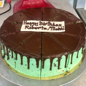 A round birthday cake with mint green frosting and a chocolate ganache top, decorated with a white fondant sign that reads 'Happy Birthday Roberta/Moht'. The cake is sliced into eight pieces.